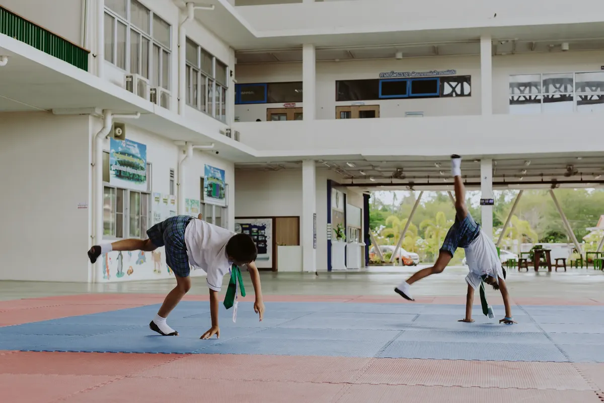 Two boys in school uniforms perform cartwheels in an indoor school gymnasium environment.