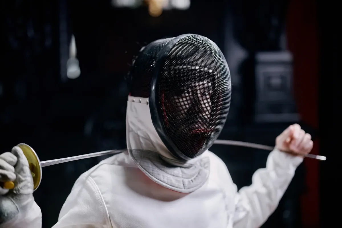 Fencer in full gear holding a foil, captured indoors with a focused expression.