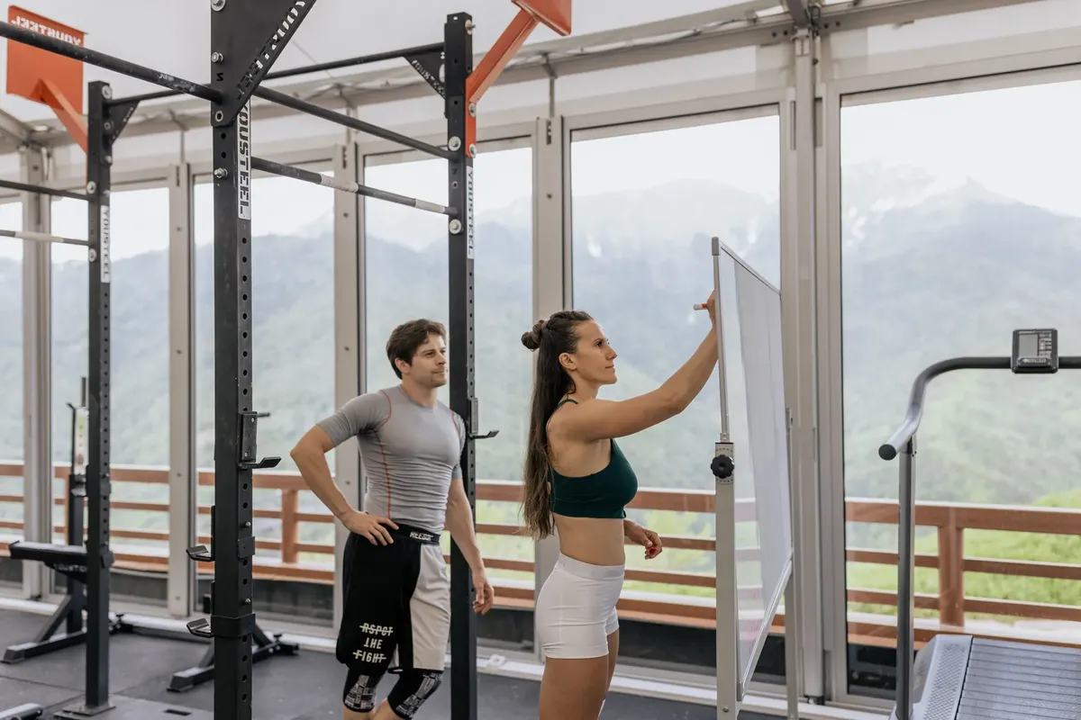 A gym coach instructs on whiteboard beside fitness equipment with mountain view.