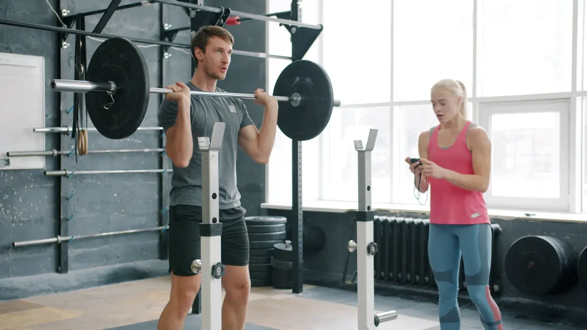 Man lifting weights while female trainer observes in a gym environment.