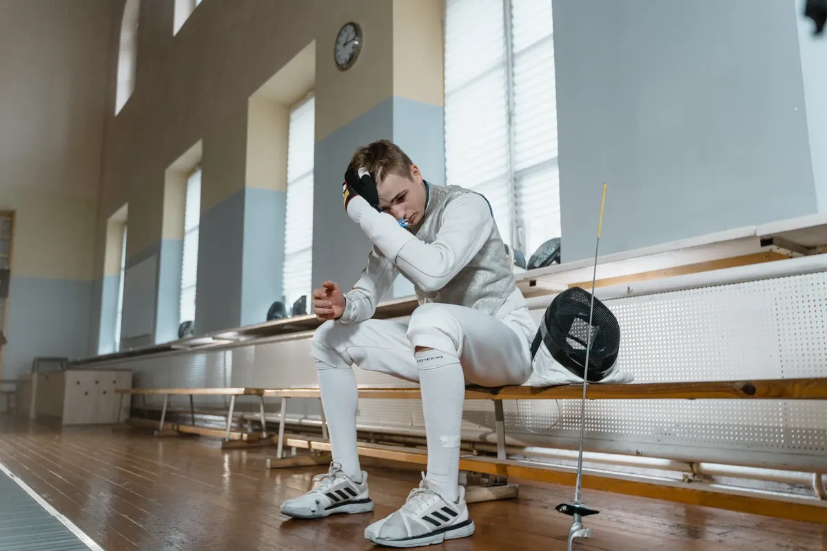 Young male fencer taking a break in a training salle, wearing full fencing gear.